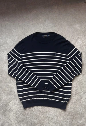 Randig tröja från Polo Ralph Lauren - Mörkblå långärmad tröja från Polo Ralph Lauren i mjuk pima bomull. Tröjan har vita horisontella ränder och en broderad logga på bröstet. Klassisk rund halsringning och ribbade . Perfekt för en clean och stilren look.