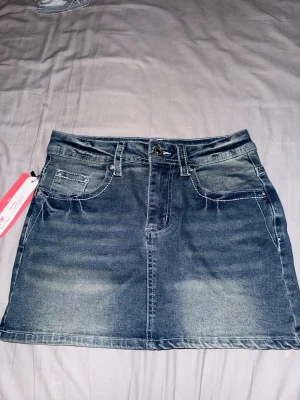 Blå jeanskjol med broderade fickor Romwe - Snygg blå jeanskjol från Romwe i storlek S. Kjolen har klassisk femficksdesign, bälteshällor och knappstängning. På bakfickorna finns detaljerade broderier med fjärilsmotiv och små pärlor. Perfekt för dig som gillar denim med extra detaljer.