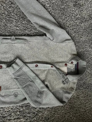 Grå kofta från Ted Baker med knappar - Snygg grå kofta från Ted Baker med mörkbruna knappar och ståkrage. Koftan är i väldigt bra skick, passar S/M