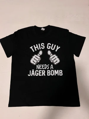 ”This Guy needs a jägerbomb” T-shirt  - En svart t-shirt med ett humoristiskt festmotiv. Trycket föreställer två pekande händer och texten “This guy needs a Jäger Bomb” i stora, tydliga bokstäver. Perfekt för partykvällar, festivaler eller som rolig present. Tröjan är i nytt skick och sitter bekvämt.