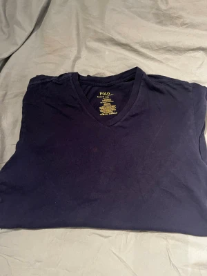 Mörkblå v-ringad t-shirt Polo Ralph Lauren - Snygg mörkblå t-shirt från Polo Ralph Lauren med v-ringad hals och klassisk vit logga broderad längst ner. Slim fit-modell i mjuk bomull, perfekt för dig som gillar stilrena och enkla plagg. Kortärmad och passar dig som vill ha en clean look.