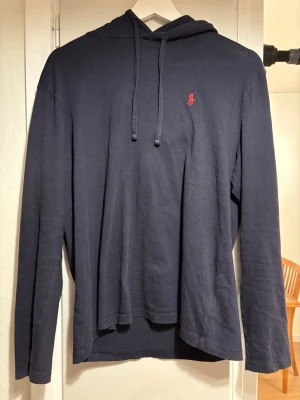 Blå Ralph Lauren Jersey Hooded T-Shirt  - Mörkblå huvtröja i mjukt material med huva och dragsnören. Broderad röd Polo Ralph Lauren-logga på bröstet. Klassisk och stilren i bra skick.