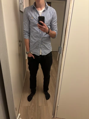 Randig skjorta från Polo Ralph Lauren - Säljer en klassisk blåvit randig skjorta från Polo Ralph Lauren med gul broderad logga på bröstet. Skjortan har lång ärm, knappar hela vägen. Perfekt för dig som gillar stilren och tidlös look.