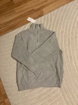 Grå stickad half zip-tröja Maison Margiela - Riktigt snygg grå ribbstickad tröja från Maison Margiela med half zip och hög krage. Perfekt för lager-på-lager och chill vibe. Långärmad och i mjukt stickat material som känns lyxigt och stilrent.
