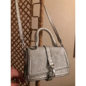 Glittrig silver axelväska från H&M - Snygg liten axelväska från H&M i glittrigt silver med både handtag och lång axelrem. Väskan har en stilren, boxig form och ett spänne framtill. Perfekt för dig som vill sticka ut med lite extra glitter och cool vibe.