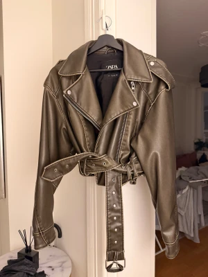 Olivgrön skinnjacka från Zara - Snygg olivgrön skinnjacka från Zara med coola detaljer som breda slag, silverfärgade dragkedjor och ett bälte i midjan. Stolek L men passar även xs/s.