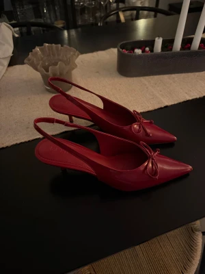 Röda pumps med rosett från NA-KD - Snygga röda pumps med slingback och spetsig tå från NA-KD. Skorna har en låg klack, elegant rosettdetalj framtill och är gjorda i glansigt syntetmaterial. Perfekta för dig som vill sticka ut med en trendig och feminin look. Nypris 799