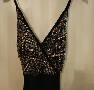 Svart långklänning med paljetter - Supersnygg svart långklänning med smala axelband och djup v-ringning. Överdelen är täckt av glittriga paljetter och pärlor i geometriskt mönster i guld och silver. Kjolen är enkel och faller rakt ner. Perfekt för dig som vill sticka ut på bal eller fest!