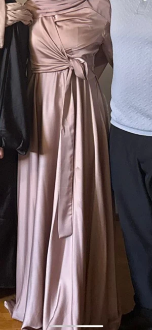 Festklänning - Säljer en elegant långklänning i champagnefärgad satin-look med långärmade ärmar och snygga manschettknappar. Klänningen har ett vackert knyt i midjan och en diskret dragkedja baktill. Flödande kjol och stilren design som ger en lyxig känsla.
