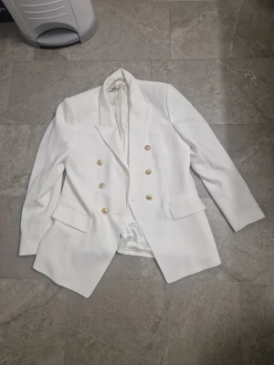 Vit kavaj från Zara med guldknappar - Snygg vit kavaj från Zara i storlek XL med dubbelknäppning och sex guldiga knappar framtill. Kavajen har klassisk krage, två fickor med lock och är helfodrad med slät insida. Perfekt för dig som vill ha en clean och stilren look. Använd fåtal gånger, någon missfärgning i kragen ( knapt att det synst) Går nog att tvätta bort. 