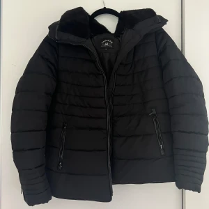 Svart pufferjacka från Crocker - Säljer en svart pufferjacka från Crocker med dragkedja och två fickor framtill. Jackan har en mysig krage med foder och är perfekt för kyligare dagar. Den är stilren och enkel att matcha med olika outfits. Storlek M. Lite av kragen har nöts bort (kolla sista två bilderna)