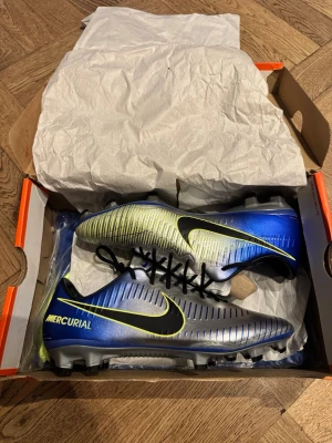 Nike Mercurial Vapor XI NJR, stl 42.5 - Säljer ett par oanvända Nike Mercurial Vapor XI (11) NJR fotbollsskor i storlek 42.5. Skorna har en snygg mix av blått, silver och neongult med svarta detaljer och snörning. Ovandelen är i syntetmaterial som snabbt formar sig efter foten, och dessutom är dem otroligt lätta. Perfekta för dig som vill sticka ut på planen. Storlek 42.5. Originalkartong samt skopåse medföljer. Otroligt eftertraktade och sällsynta, framförallt i denna storlek och skick.
