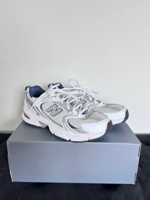 New Balance sneakers, storlek 39.5 - Säljer ett par vita New Balance sneakers med marinblå detaljer. Skorna har en sportig design med meshdetaljer för extra ventilation, snörning och en robust platt sula. Perfekta för dig som gillar retrostil. Ikoniskt N på sidan och vadderad häl för skön passform. Sparsamt använda 👟✅ Avtvättade så gott det går - skosnören är t.o.m tvättade i maskin. Kommer i originalförpackning. Önskar du veta exakta mått? Tveka inte att höra av dig!