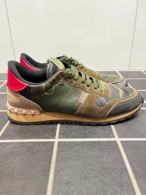 Valentino Rockrunner  - Snygga Valentino Rockrunner sneakers med camouflage-mönster i grönt, brunt och svart. Skorna har detaljer i mocka och skinn, snörning och en beige sula med nitar samt röd häl. Perfekt för dig som gillar streetstyle med lyxig touch.