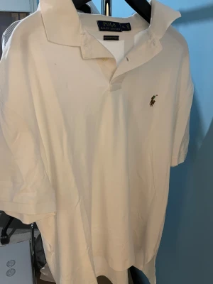 Vit piké från Polo Ralph Lauren XL - Vit klassisk pikétröja från Polo Ralph Lauren i storlek XL. Mjuk bomullskvalitet med Pima Soft Touch, korta ärmar och krage med knappslå. Ikonisk broderad logga på bröstet. Perfekt för en clean och stilren look.