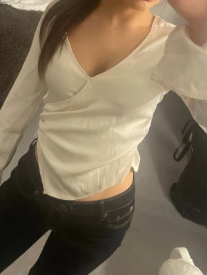 Vit omlottopp med v-ringning - Säljer en stilren vit omlottopp med v-ringning och långa ärmar. Toppen har en croppad passform och är tillverkad i ett mjukt, glansigt material som ger en lyxig känsla. 