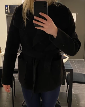 Svart omlott kappa med bälte - Snygg svart kappa med bred krage och omlott-design. Jackan har ett matchande bälte i midjan och två stora fickor framtill. Perfekt för dig som gillar stilrena och klassiska plagg med en modern twist. Passar dig som vill ha en jacka med lite längre längd och enkel look. Storlek M men passar S.