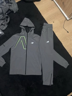 Grå New Balance träningsset - Säljer ett grått träningsset från New Balance med matchande hoodie och byxor. Setet har vita NB-loggor och en grön grafisk detalj på jackan. Byxorna har dragsko i midjan och dragkedja vid benslut. Perfekt för dig som gillar sportig och clean stil. XS-XL