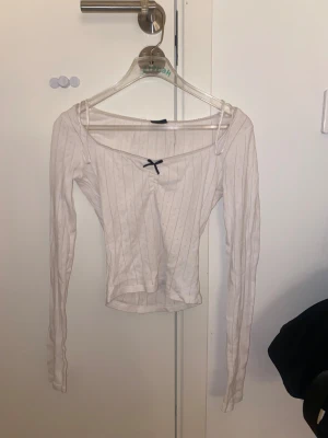 Vit långärmad topp med rosett Gina Tricot - Supersöt vit långärmad topp från Gina Tricot i storlek XS. Toppen har ett diskret hålmönster med smala ränder, v-ringad hals med liten svart rosett och spetskant. Materialet är mjukt och stretchigt, perfekt för dig som gillar en feminin och chill stil.