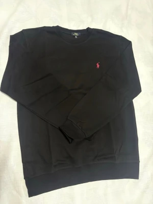 Ralph Lauren Tröja - Svart sweatshirt från Polo Ralph Lauren med klassisk rund hals och lång ärm. Tröjan har en liten rosa broderad logga på bröstet och ribbade muddar vid ärmslut och nederkant. Perfekt för en chill och stilren look.