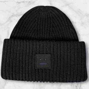 Svart stickad mössa Acne Studios - Svart ribbstickad mössa från Acne Studios med klassisk face-logga framtill. Tillverkad i 100% ull för extra värme och mjuk känsla. Perfekt accessoar för kyliga dagar och enkel att matcha med olika stilar.