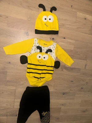Gul och svart bi-dräkt - Söt väst för bebis i gult och svart med bi-tema. Västen har randigt mönster, broderade ögon och antenner samt små vingar på sidorna. Perfekt för dig som vill ha något lekfullt och gulligt till din lilla.Sött set med en gul body med svart randigt bi-tryck och matchande svarta byxor med ett bi-tryck på benet. Setet inkluderar även en gul mössa med bi-design. Halloween 🎃 eller annat tema