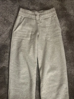 Ljusgrå mjukisbyxor XS med snörning - Ljusgrå mjukisbyxor från 157 i storlek XS. Byxorna har bred resår i midjan med snörning och två stora fickor framtill. Raka ben och mjukt material som känns skönt mot huden. Liten defekt på framsida lår men som ej syns mycket.