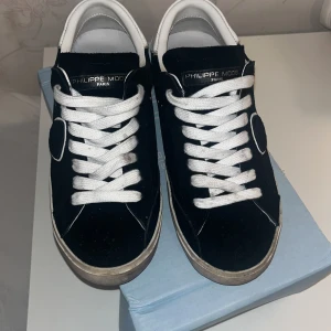 Philippe Model svarta sneakers mocka - Svarta Philippe Model sneakers i mocka med vita skinn-detaljer och vit platt sula. Klassisk låg modell med snörning och rund tå. Snyggt sköldmärke på sidan och metallnitar för extra edge. Perfekt för dig som gillar stilrena och trendiga sneakers.