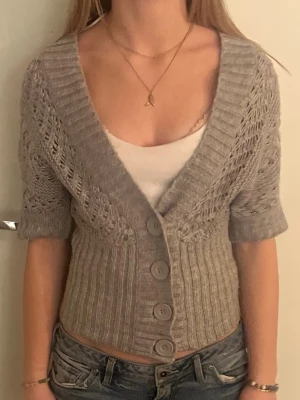 Grå stickad kofta med kort ärm - Snygg grå stickad kofta med korta ärmar och stora knappar framtill. Djup v-ringning och ribbstickad midja ger en schysst passform. Koftan har ett fint hålmönster på ärmarna och upptill. Perfekt att slänga över ett linne. Inköpt för 300 och inte använd av mig så lägger mig på 250, men priset kan diskuteras🫰🏻🩷