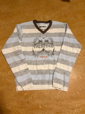 Randig stickad tröja Calavera - Stickad tröja med breda ränder i ljusblått, grått och vitt från Calavera. V-ringad hals med brun ribbad kant och tryckt motiv samt text på bröstet. Långa ärmar och mjukt material, perfekt för en avslappnad stil. Mått: längd: 65cm bredd: 50cm armar: 65cm 