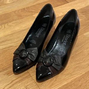 Svarta pumps med rosett från Starlet - Snygga svarta pumps från Starlet med spetsig tå och dekorativ rosett framtill. Skorna har en elegant låg klack och är tillverkade i glansigt syntetmaterial. Storlek 4 1/5 och passar mig som har storlek 37