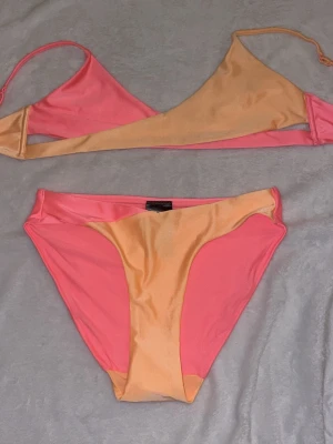 Tvåfärgad bikini i rosa och persika färg - Säljer en snygg bikini med trekantsöverdel och klassiska bikinitrosor. Båda delarna är tvåfärgade i rosa och orange/persika färg, vilket ger en cool och trendig lock. Perfekt för strand eller pool. Inga vadderingar i toppen. Underdelen är inte använd har lappen kvar. 
