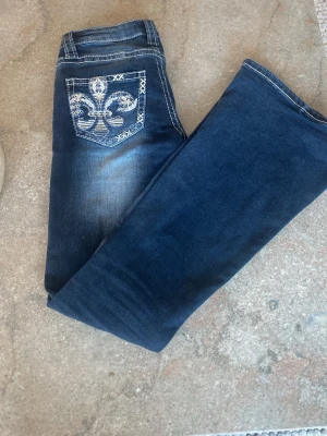 Mörkblå jeans med broderad bakficka - Snygga mörkblå jeans med broderad bakficka i vitt och silver. Jeansen har klassisk femficksmodell, kontrastsömmar och en unik detalj på bakfickan. Perfekta för dig som vill sticka ut lite extra med dina jeans.