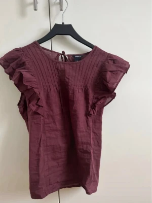 Vinröd blus med volang från Lindex - Supersöt vinröd blus från Lindex med volangdetaljer på ärmarna och knytning i nacken. Blusen har rund halsringning och är tillverkad i ett lätt bomullstyg. 