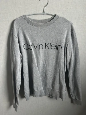 Grå sweatshirt från Calvin Klein - Snygg grå sweatshirt från Calvin Klein med stor logga tryckt över bröstet. Tröjan har rund halsringning, långa ärmar och är tillverkad i mjukt sweatshirtmaterial. Perfekt för en avslappnad och trendig look.