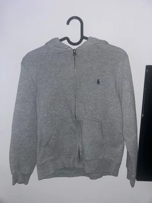 Grå zip hoodie från Polo Ralph Lauren - Klassisk grå zip hoodie från Polo Ralph Lauren med liten mörkblå logga broderad på bröstet. Hoodien har huva, dragkedja framtill och två stora fickor. Tillverkad i mjukt sweatshirtmaterial och har ribbade muddar vid ärmar och nederkant.