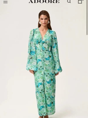 Blommig långklänning från Adoore - Snygg långklänning från Adoore i grönt och blått blommigt mönster. Klänningen har v-ringning, långa vida ärmar och knappar framtill. Perfekt för dig som vill sticka ut med en färgglad och feminin look. En knapp lossnat.