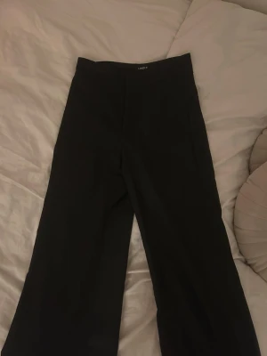 Svarta kostymbyxor från Lindex - Stilrena svarta kostymbyxor från Lindex, modell Lykke. Byxorna har en klassisk rak passform och hög midja. I storlek 36
