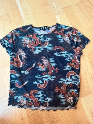 Mesh topp med drakmönster från iStay - Snygg mesh topp från iStay med asiatiskt inspirerat drakmönster i rött och vitt mot en mörkblå bakgrund. Toppen är kortärmad och har vågiga kanter nedtill och vid ärmsluten. Perfekt för dig som vill sticka ut med ett coolt print.