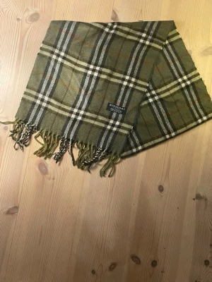 Grön rutig halsduk från Burberry - Snygg grön halsduk från Burberry med klassiskt rutmönster i beige, svart och vitt. Halsduken har fransar i ändarna och en diskret Burberry-logga. Perfekt accessoar för att lyfta din outfit med en tidlös touch. Pris kan diskuteras.