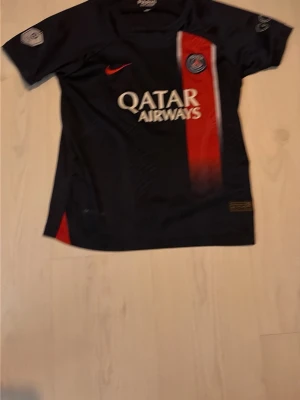 PSG fotbollströja Mbappé #7 - Mörkblå PSG matchtröja från Nike med röd och vit rand, Qatar Airways-tryck på bröstet och Mbappé #7 på ryggen. Tröjan har korta ärmar och klubbmärke på bröstet. Perfekt för fotbollsfans!