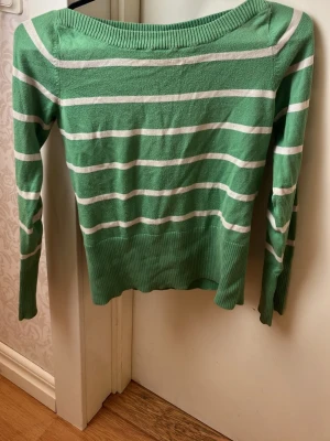 Grön och vit randig tröja - Grön långärmad tröja med vita horisontella ränder och bred ribbad mudd vid ärmslut och nederkant. Tröjan har en klassisk rund halsringning och är tillverkad i ett mjukt, stickat material. Perfekt för en avslappnad och stilren look.