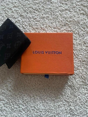 Svart plånbok i skin  - Snygg svart plånbok med klassiskt monogrammönster. Tillverkad i exklusivt skinn och levereras i original orange box. Perfekt för dig som vill ha en stilren och lyxig accessoar i fickan.