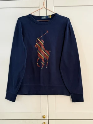 Mörkblå sweatshirt Polo Ralph Lauren - Mörkblå sweatshirt från Polo Ralph Lauren med stort färgglatt polospel-tryck på bröstet. Tröjan har rund halsringning och ribbade muddar vid ärmar och nederkant. Tillverkad i mjukt bomullsmaterial, perfekt för en avslappnad stil.