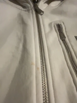 Vit hoodie från Sail Racing - Vit hoodie från Sail Racing med dragkedja framtill och en extra dragkedjeficka på bröstet. Hoodien har broderad logga på bröstet och är tillverkad i mjukt material. Perfekt för en avslappnad och sportig stil.