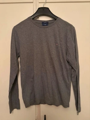 Grå långärmad tröja från Gant - En stilren grå långärmad tröja från Gant i mjuk bomull. Tröjan har rund halsringning och en liten broderad Gant-logga på bröstet. Aldrlg använd!