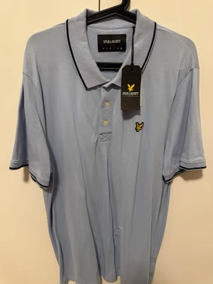 Ljusblå pikétröja från Lyle & Scott - Snygg ljusblå pikétröja från Lyle & Scott med klassisk krage, knäppning med tre knappar och svart piping på krage och ärmslut. Tröjan har den ikoniska gula loggan broderad på bröstet och är tillverkad i ett mjukt pikématerial.