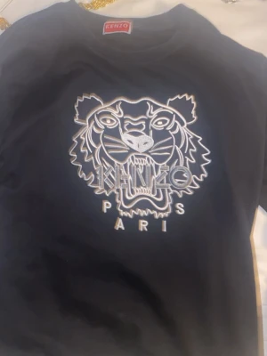 Svart sweatshirt från Kenzo med tiger - Cool svart sweatshirt från Kenzo med det ikoniska tigermotivet och texten 'KENZO PARIS' broderat i vitt på bröstet. Tröjan har rund halsringning och ribbade muddar. Perfekt för dig som vill sticka ut med en streetwear vibe.