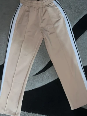 Beige mjukisbyxor Palm Angels - Snygga beige mjukisbyxor från Palm Angels med vita och svarta ränder längs sidorna. Byxorna har resår i midjan, raka ben och sidofickor. Palm Angels-loggan är tryckt på vänster sida. Perfekta för en chill och trendig look.