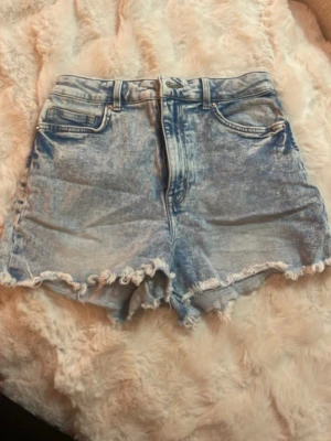 Blå högmidjade jeansshorts - Säljer ett par blå högmidjade jeansshorts med fransig kant och klassisk femficksdesign. Shortsen har en ljus effekt och råa kanter för en avslappnad look. Perfekta för varma dagar och enkel att matcha med olika toppar.